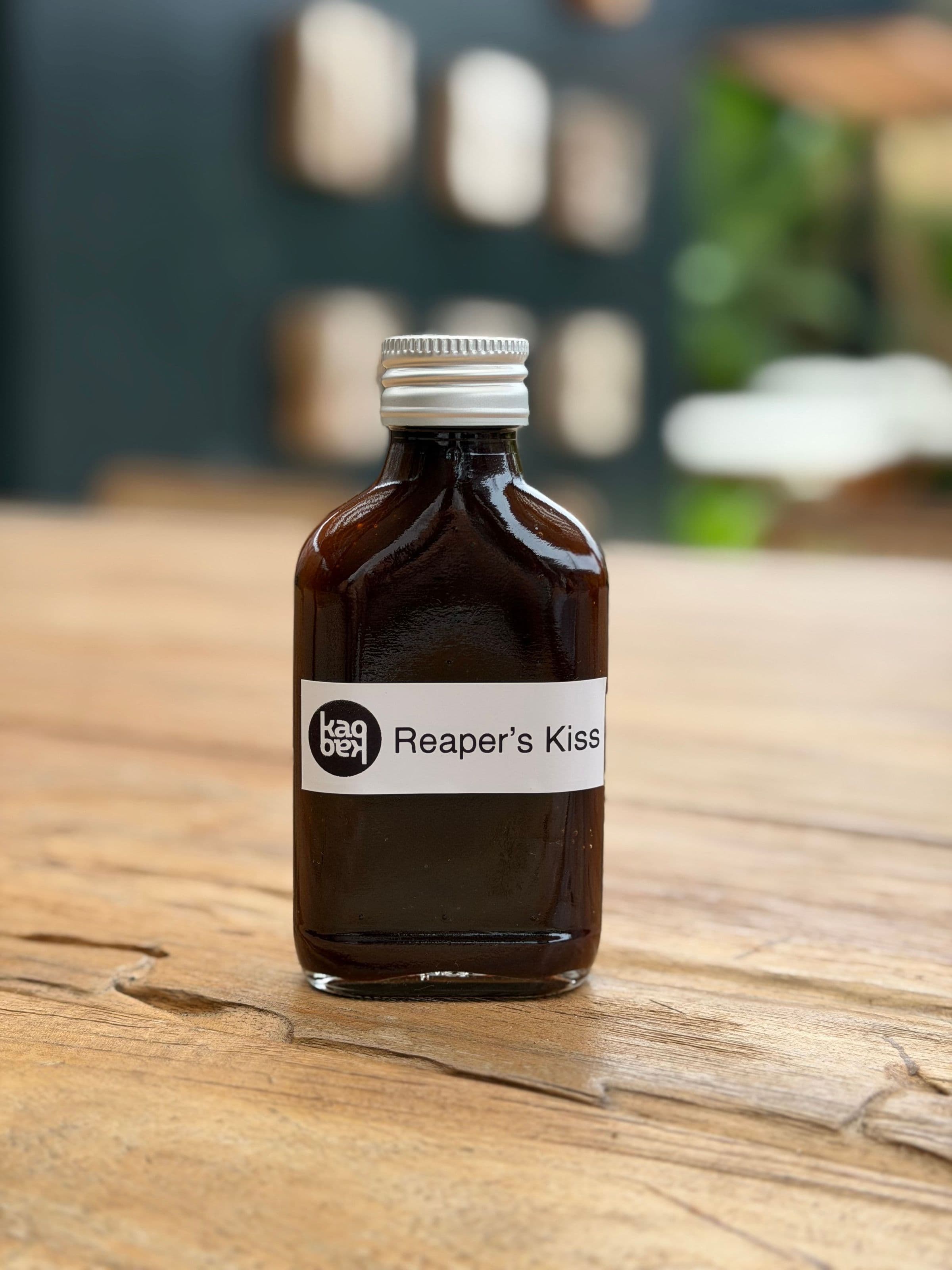 Reaper’s Kiss hot sauce in a dark, amber-brown glass bottle with a silver screw cap and minimalist white label, standing on a rustic wooden table with a muted, blurred background.